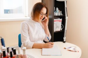 A blonde woman in glasses and a beautician outfit, talking on the phone and writing something in a notebook.