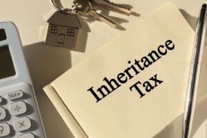 Notepad with the phrase Inheritance Tax, calculator and keys with house shaped key chain on white table.