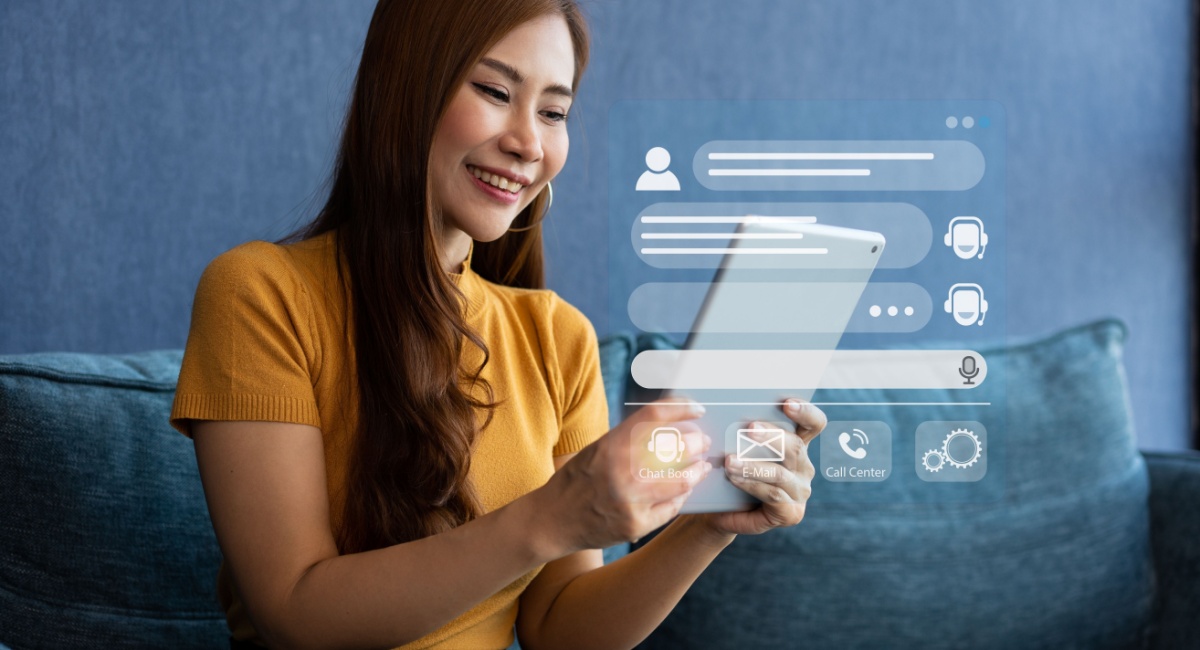 A woman holding a tablet and smiling as she engages in a positive customer experience via online chat. Icons appear over the tablet to indicate messages being exchanged between the customer and customer care agent